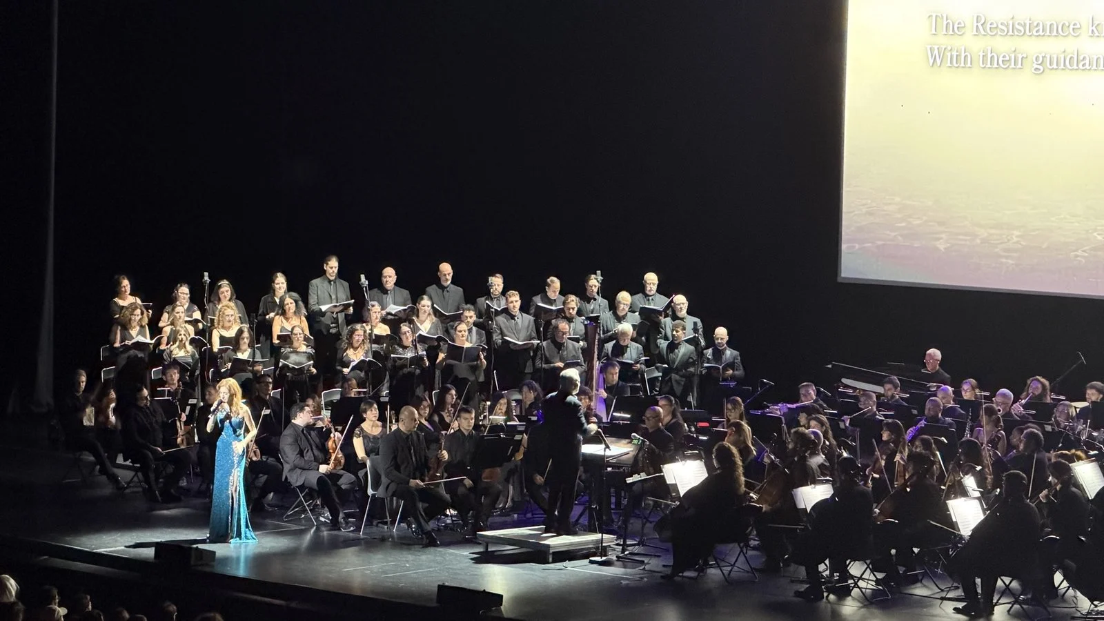 L'orchestre et le chœur sur scène, avec le récit sous-titré en anglais projeté sur l'écran